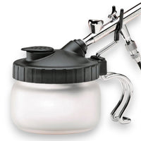 A white cleaning station can with a black ribbed screw-on lid, shown with a silver airbrush connected to the top, on a plain white background.